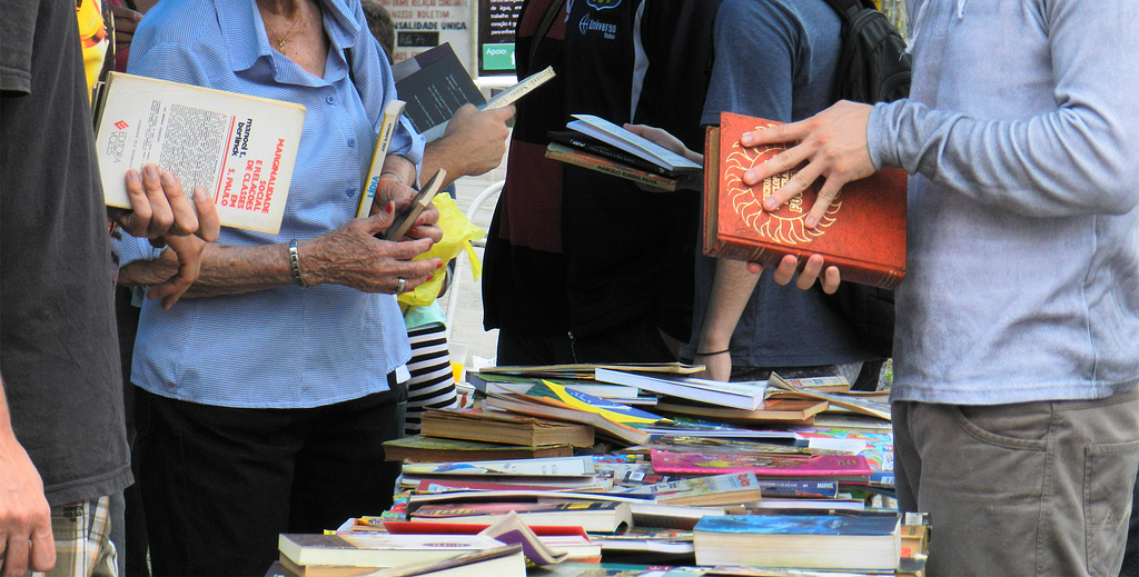 Close em mesa coberta por livros. Grupo de pessoas (adolescentes, jovens adultos e idosa) garimpam o livros.