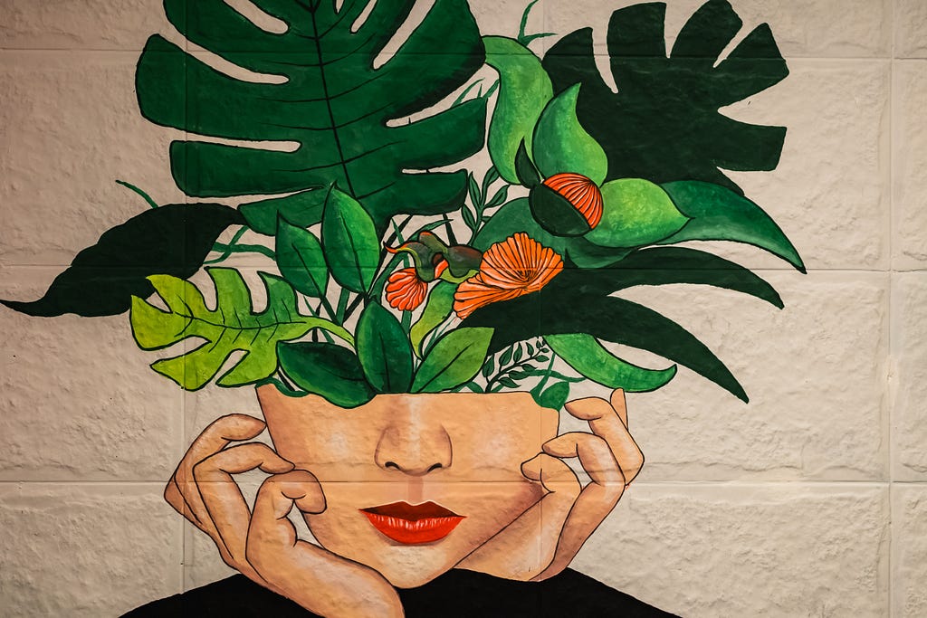 A painting of half a woman’s face on a white wall with her fingers curled around her cheeks. Ferns are growing out of her head, symbolic of growth.