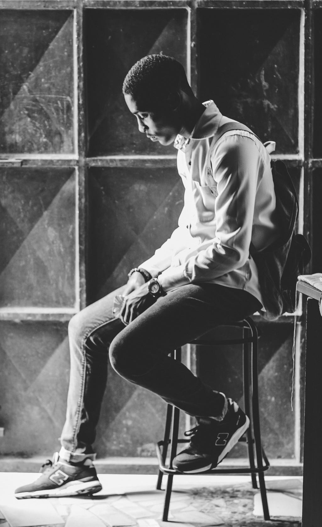 A gray-scale picture of male teenager sitting on a stool. His hair is short, black, and wavy. His skin is brown. He is wearing a light-colored shirt, denim pants, and a dark, empty backpack.