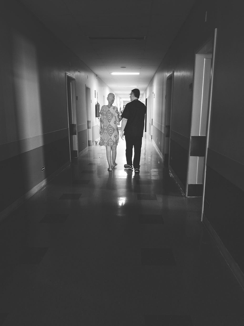 Foto em preto e branco da minha mãe e o fisioterapeuta de costas, caminhando no corredor do hospital.
