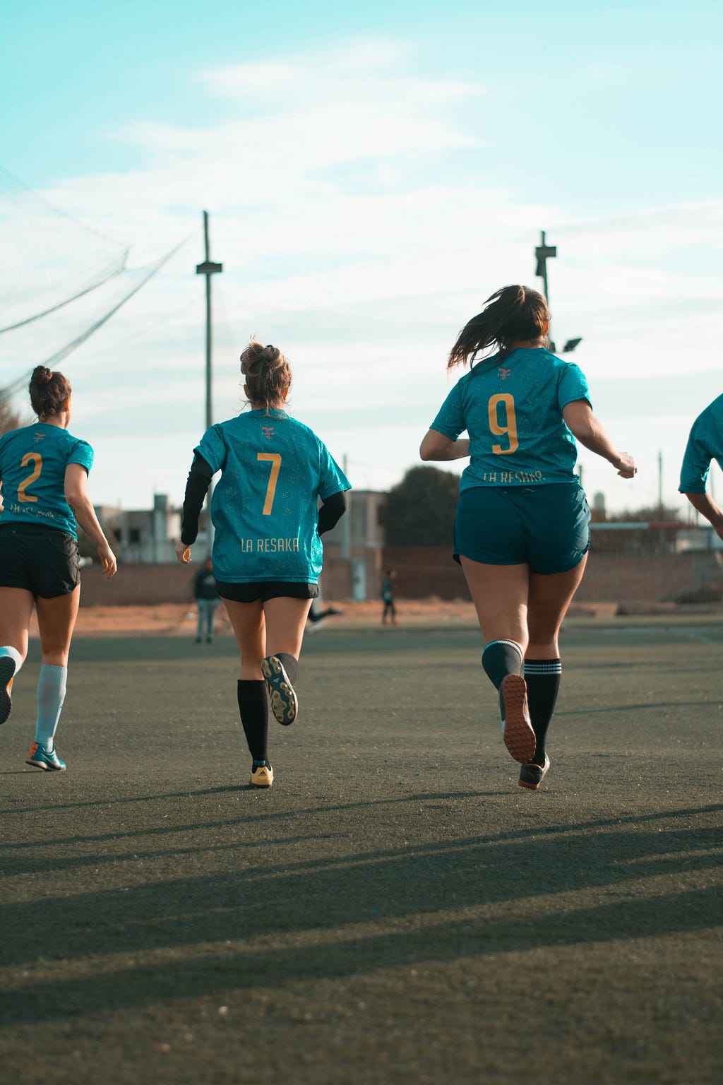 A group of three women wearing teal sports jerseys running on a green field