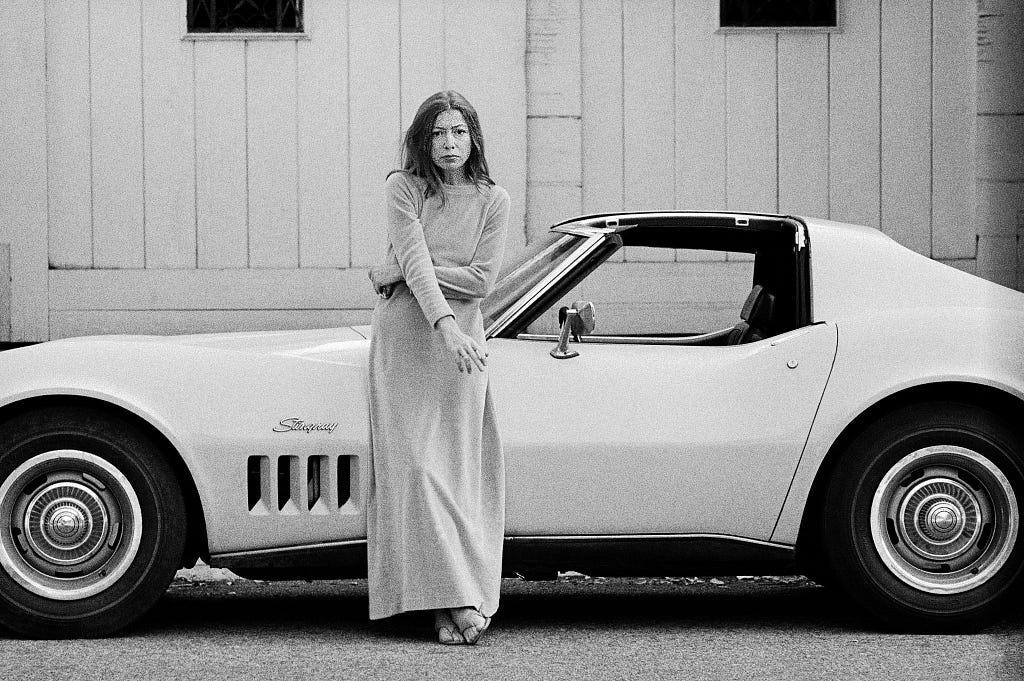 Escritora Joan Didion, uma mulher branca de cabelos escuros e um vestido claro, apoiada à lateral de um Corvette. É uma foto preto e branco e Didion segura um cigarro na mão direita