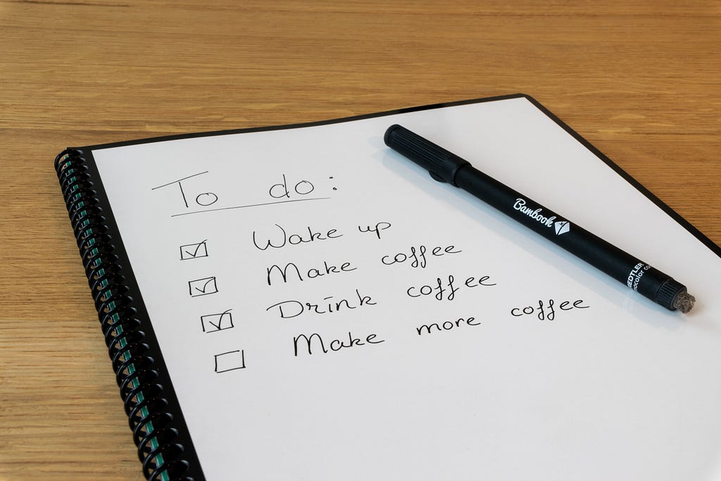 A checklist on a notepad with a pen, reading “Wake up”, “Make coffee”, “Drink coffee”, “Make more coffee”. The first three items are ticked.