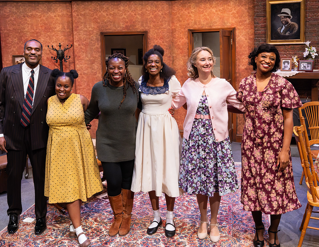 In a post-show curtain call, five actors dressed in 1950s costumes stand with another woman (their director) in modern-day attire; all smile toward the camera.