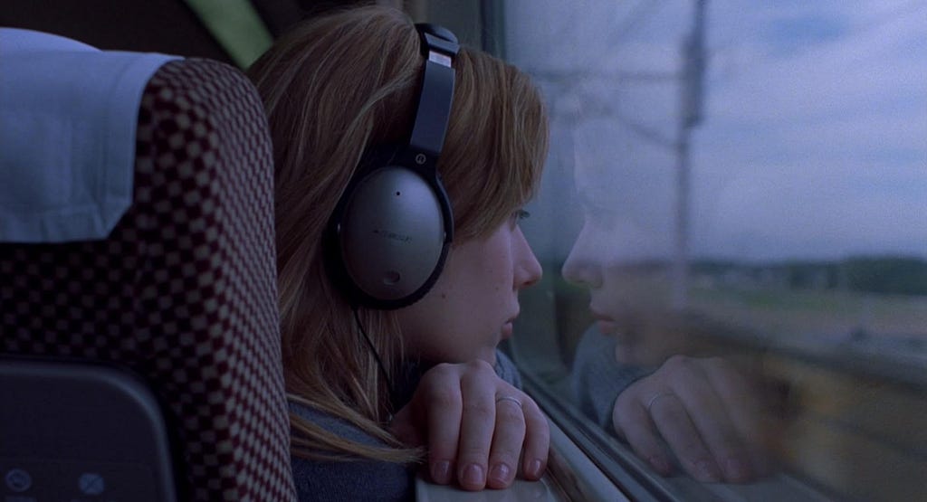 Scarlett Johansson, a blonde woman wearing large headphones and looking out the window of a vehicle with a focused expression; blue light in the background and an outdoor setting. A scene from the movie Lost in Translation.