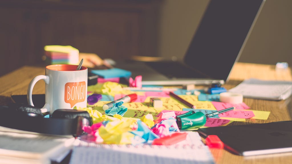 A messy desk with lots of colourful post it notes, a laptop and a mug. (Not my desk) Photo: Ferenc Horvath on Unsplash
