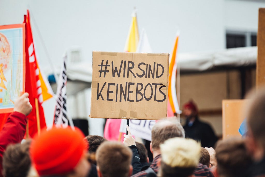 In a crowd, a cardboard sign is held up saying #WirSindKeineBots in German, which means — We are not robots in English
