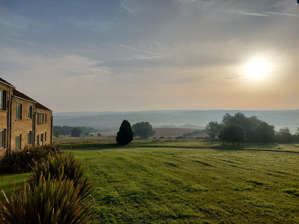 Sun rising over County Durham. A rough lawn of grass and reeds, with trees and rolling hills in the background. A stone hotel building, 2 stories high, is down the left side of the picture.