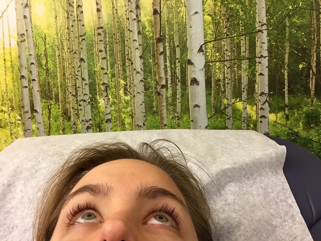 Woman lying on doctor’s table, staring at the ceiling. Behind her is birch tree wallpaper.