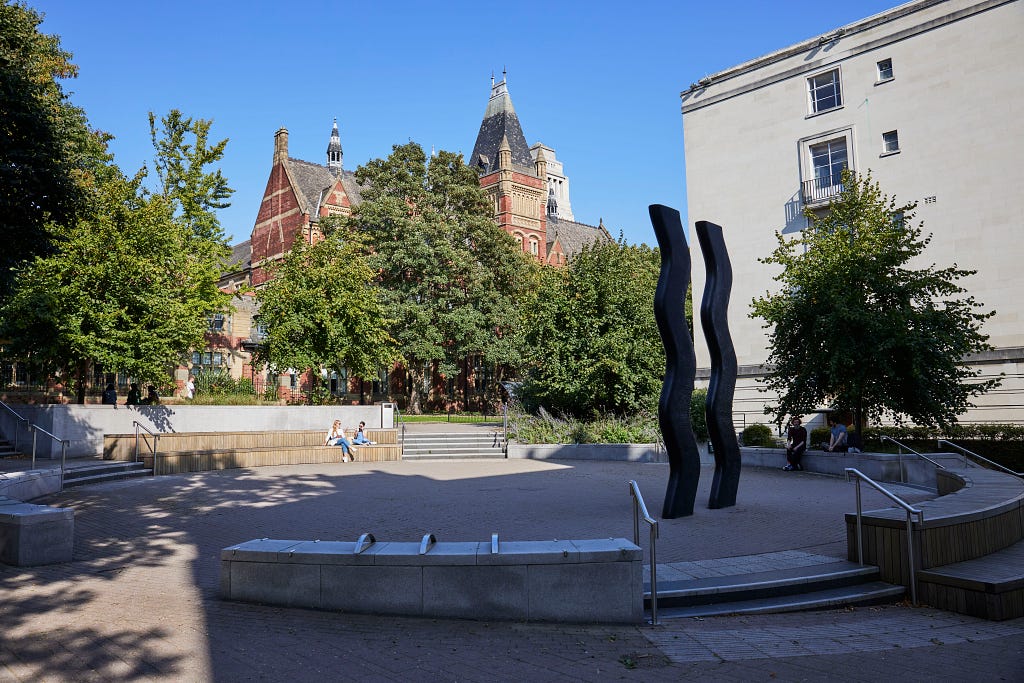 The plaza is shaded by large green trees on an otherwise sunny day. The sign for art statue consists of two large, separate wavy lines, made of thick black metal.
