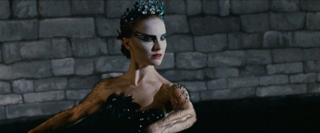 The image shows the actress wearing a black gown, dancing ballet. Her eyes have sharp black makeup and she has a big tiara on her head. Her arms have little feathers sprouting through her skin.