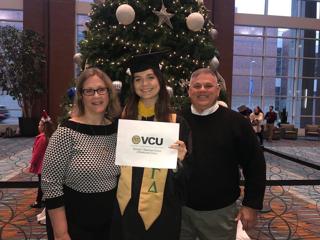 My mom, myself in a graduation stole and cap, and my dad standing in front of a Christmas tree. I am folding my VCU Graduation folder.