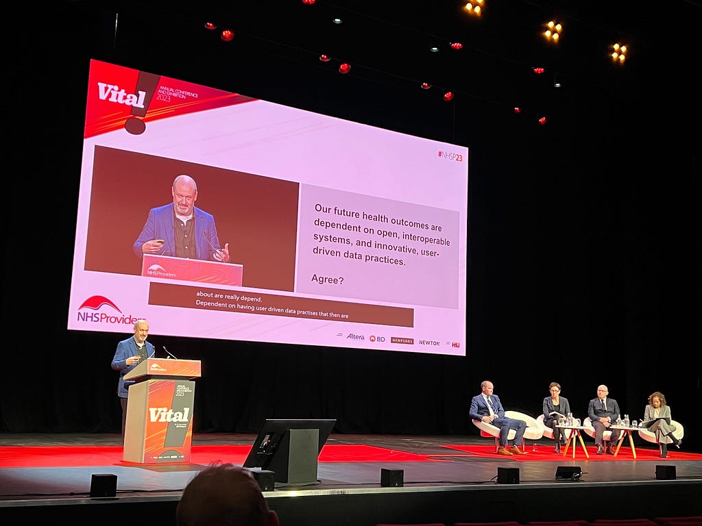 Mike Bracken founding partner of Public Digital talking at a lecturn at the NHS Providers conference. On the screen in the background is a slide that says “Our future health outcomes are dependent on open, interoperable systems, and innovative, user-driven data practices. Agree?”