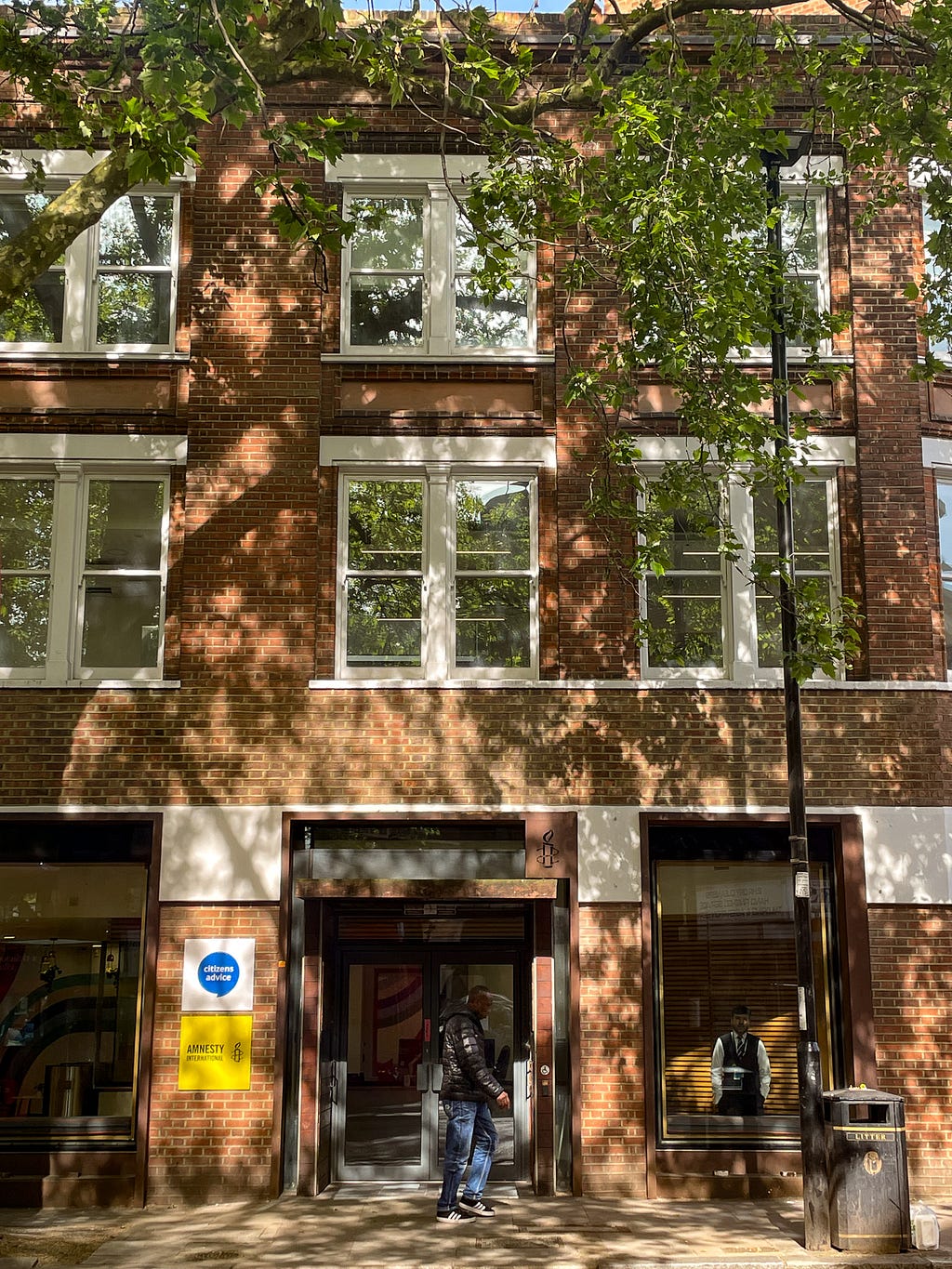 The front of a redbrick office building in London, with trees and the shadows from the trees. Next to the front door, there are logos for Citizens Advice and Amnesty International.