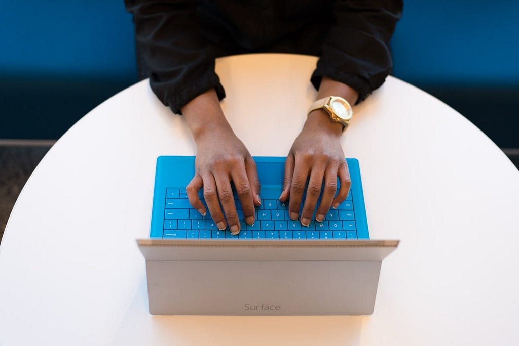 A photo taken from above shows a person’s hands typing on the blue keyboard of a laptop. The person is wearing a dark colored long sleeved shirt and a watch. The laptop is sitting on a round white table. Only a portion of the table is captured in the photo.