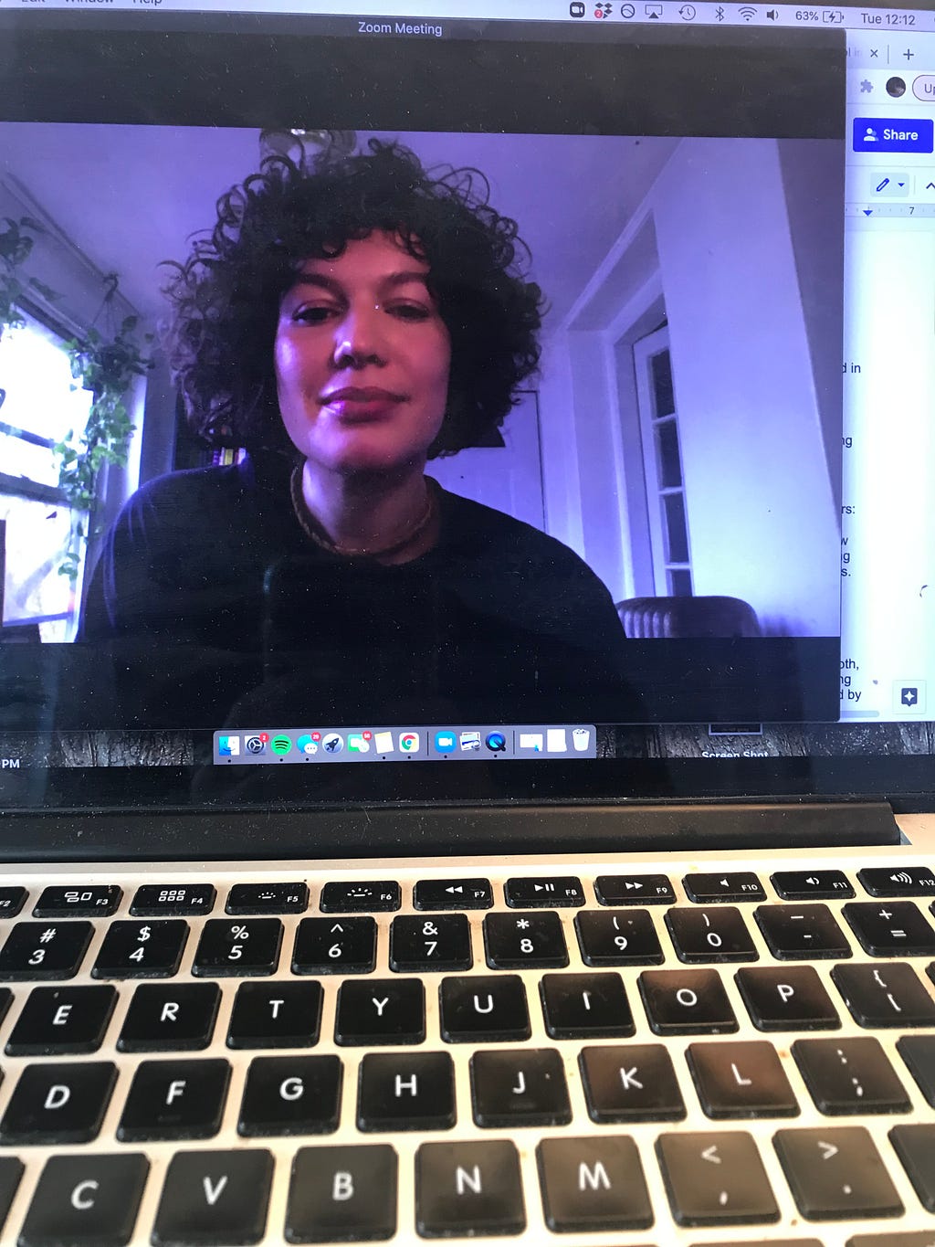 A macbook with the screen displaying a web browser with multiple tabs open in the background. In the foreground of the image is Coralys Carter, light-skinned with chin length dark brown hair and a black t-shirt with a thick gold chain, waiting to enter a Zoom meeting.