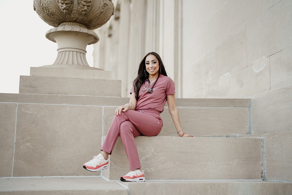 Image of Lauren Waddell, a nurse practitioner, wearing mauve scrubs and a stethoscope. She is seated with her legs crossed on large stone steps.