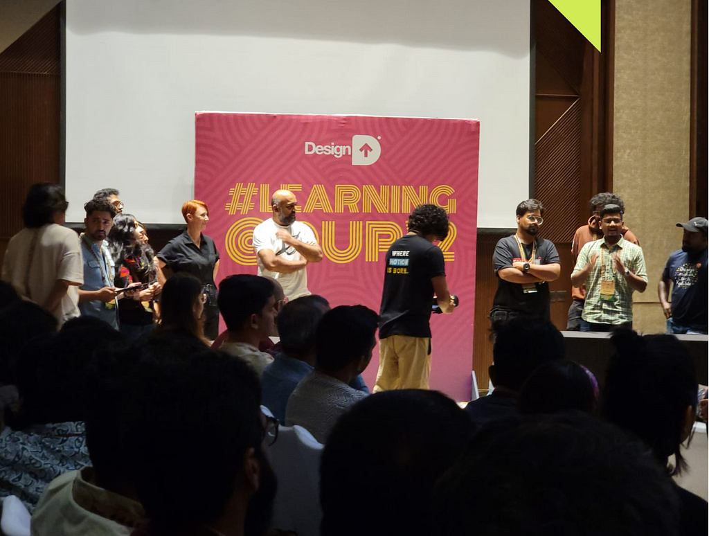 People discussing a topic in the backdrop of a DesignUp poster with a few people on stage and in limelight.