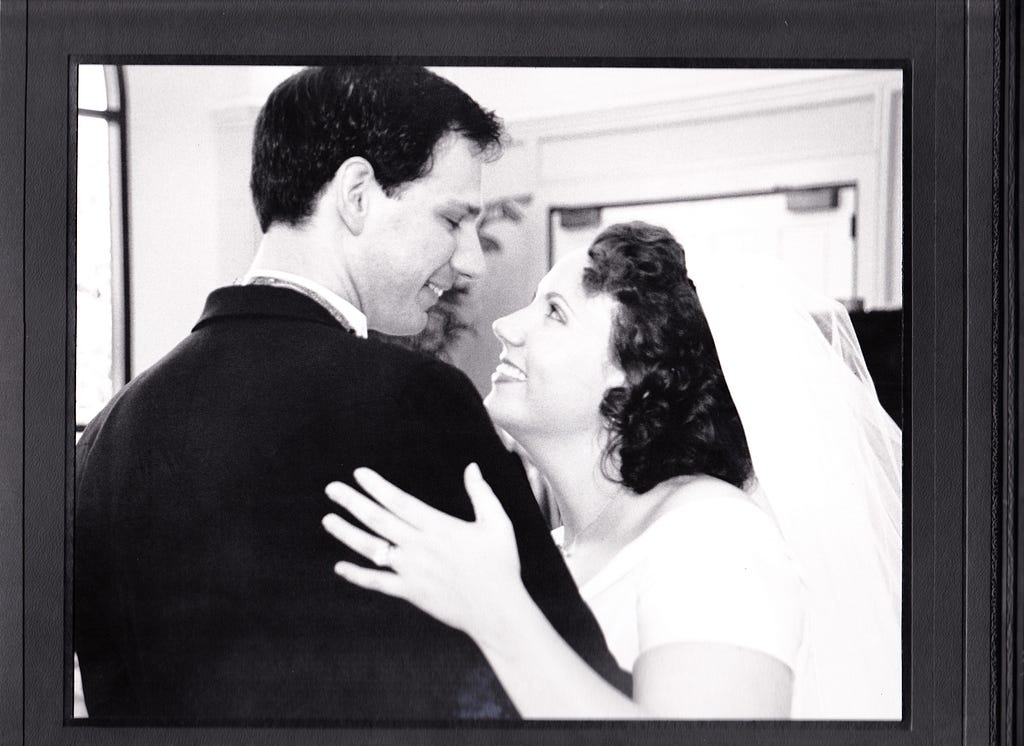 A groom dressed in tuxedo and bride dressed in white gown and veil smile while looking into one another’s eyes as they dance.