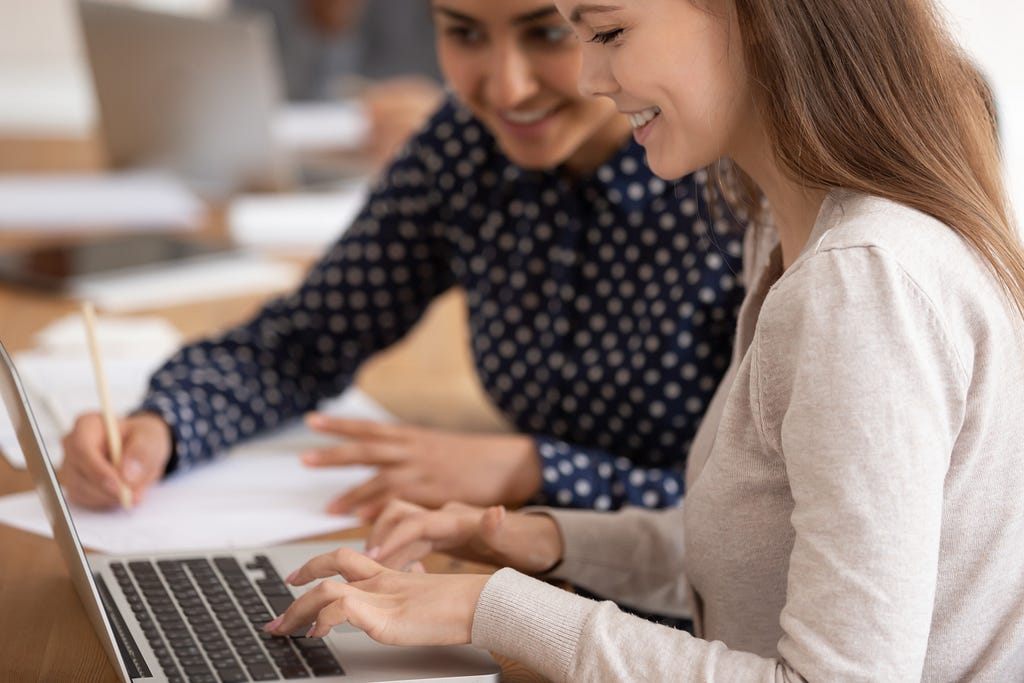 2 women sat by a computer, one using the computer and one making notes.