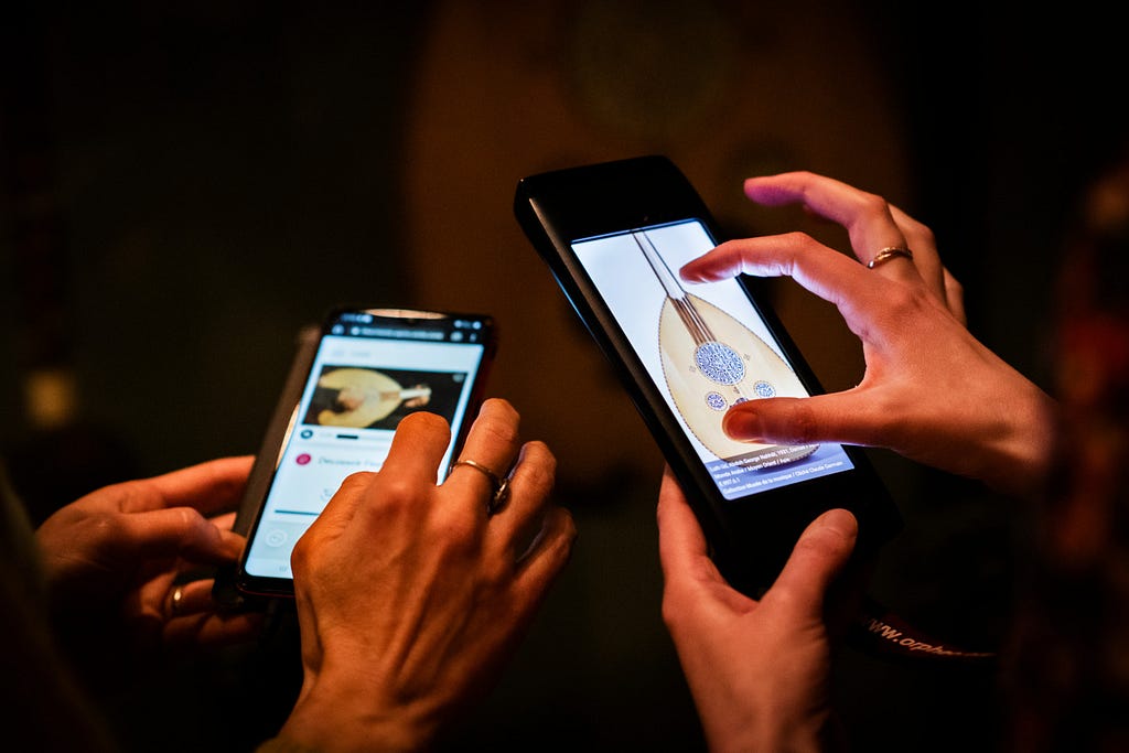 “Two hands hold smartphones, one showcasing an app with a traditional stringed instrument, the other blurred in the background, enhancing an interactive learning experience in a dimly lit environment.”