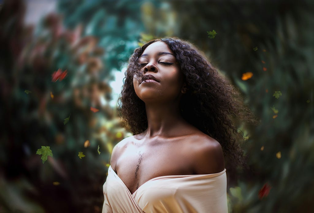 A photo of a Black woman in a cream, off the shoulder dress who’s looking up with her eyes closed by with an expression of peace and strength.