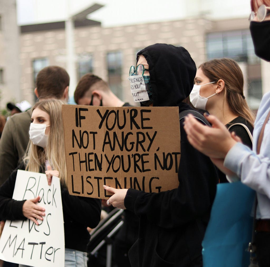 A woman dressed entirely in black in the middle of a protest, holding a sign that reads: “If you’re not angry, then you’re not listening.” She has another sign placed over her cloth mask, which partially reads: “Silence makes you complicit.”