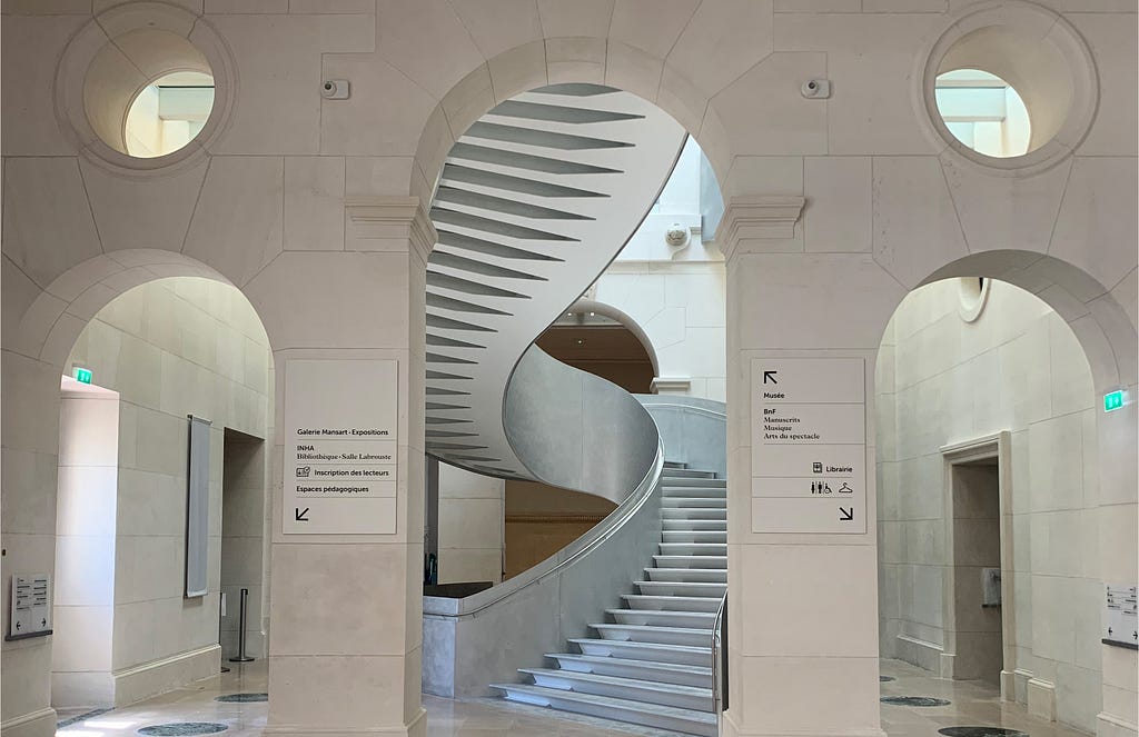 BNF Richelieu, a modern, elegant spiral staircase stands out in the center of a bright, spacious hall, with light-colored stone walls and arches. Signposts guide you to various cultural areas such as the gallery, library and museum.