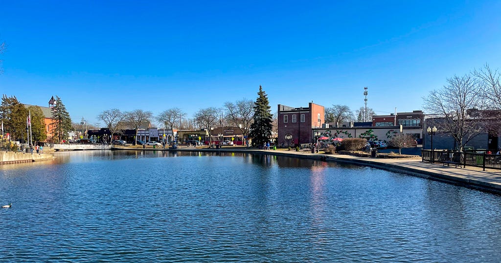 Across a millpond there is a street of buildings and trees.