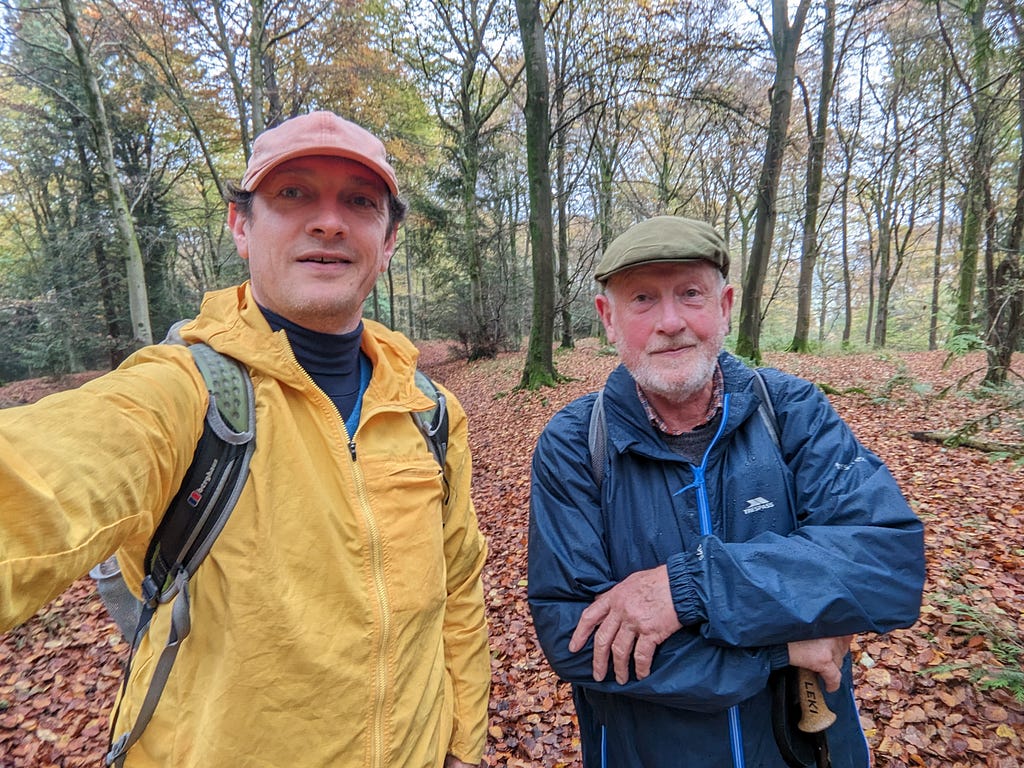 Two men (my dad and me) wearing waterproof coats and hats in a wood along the route of the South Downs Way. It is autumn and the ground is covered with red and brown leaves.