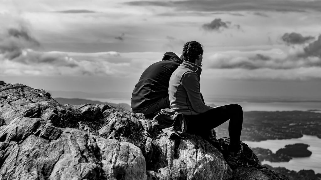 A grayscale photo of two people sitting on mountain rock, overlooking scenery below/ahead. They wear winter hiking gear.