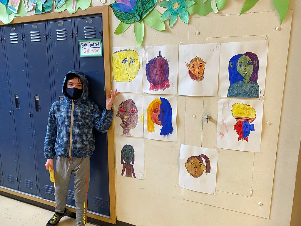A kid is gesturing to his drawing hung with several drawings by other kids in a school hallway.