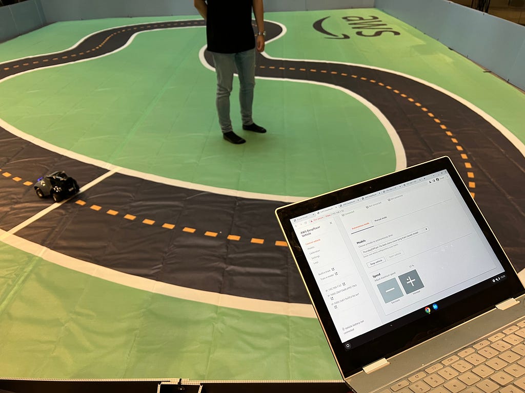 Laptop in the foreground of the miniature-sized race track with a remote control car driving past the finish line.