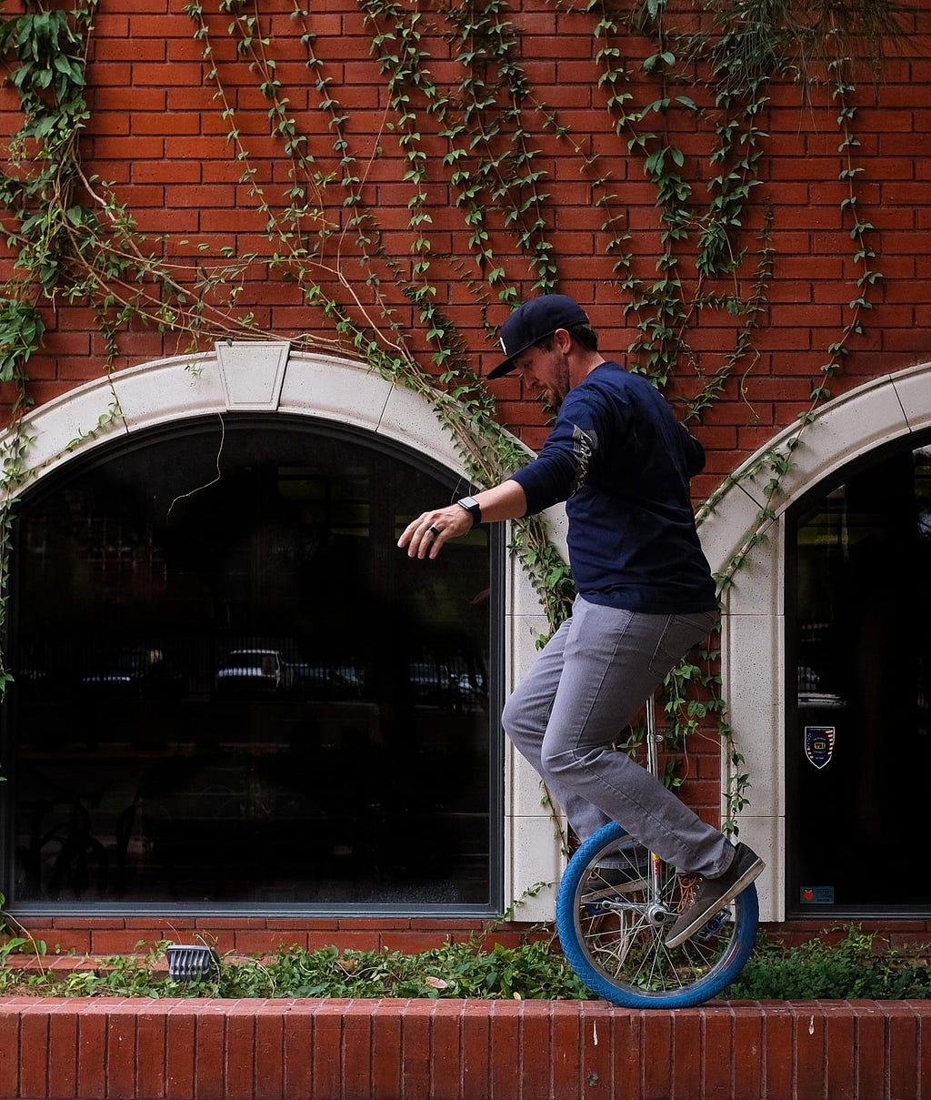 A man rides a blue unicycle atop a low brick wall