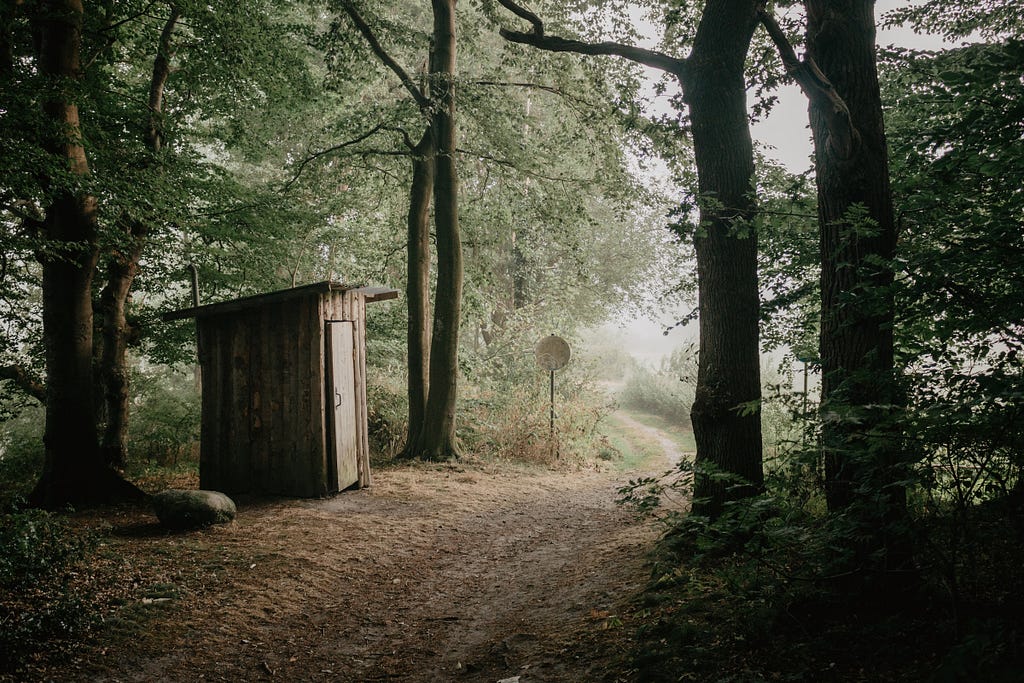 A forest path with a wooden outdoor restroom on the left