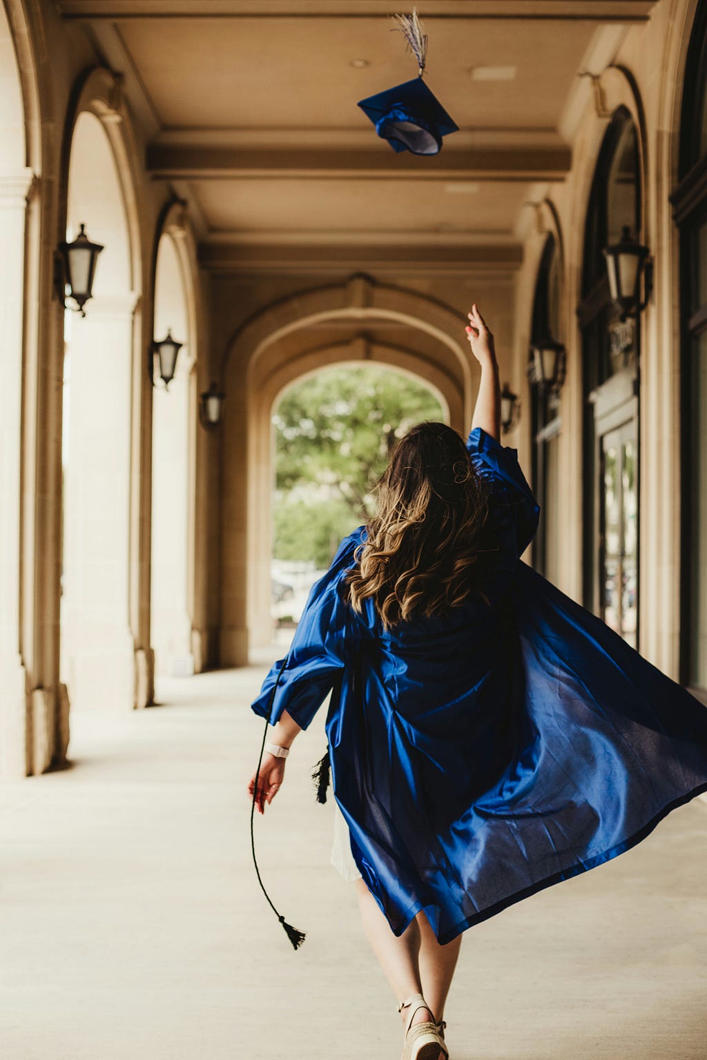 woman in a blue graduation gown throwing her graduation cap up in the air while walking