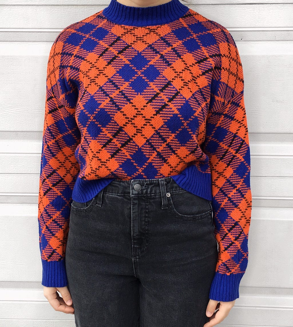 An orange turtle neck sweater with a purple diamond and crisscross design. Worn with high-waisted grey jeans.