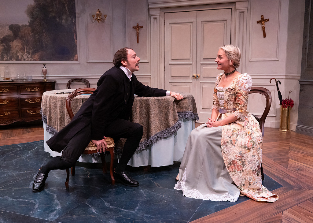 A man wearing all black 19th century clothes sits and leans lustfully toward a smiling woman in a floral lace early 1900s gown, who smiles politely.