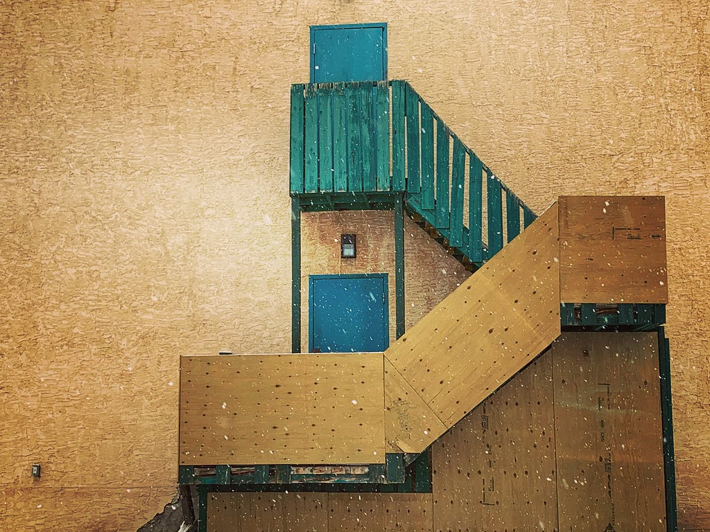 Side of an old building in brownish particle board with a wooden staircase leading to a green staircase and a blue door.