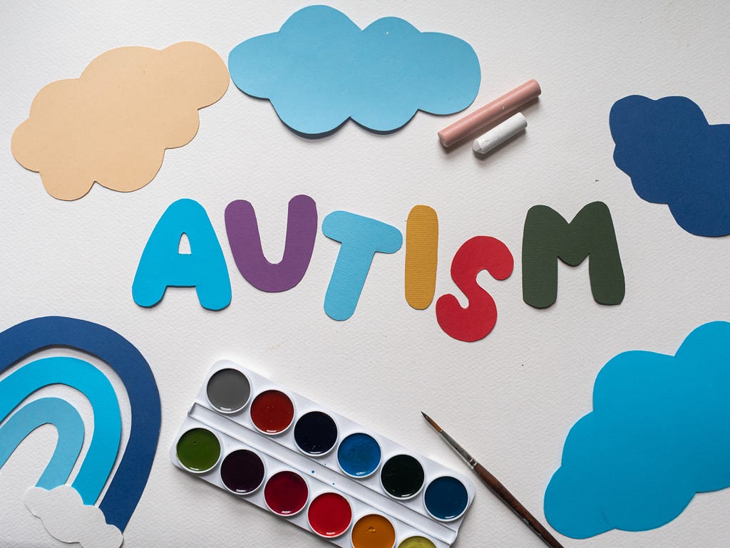 The word AUTISM. Each letter is cut from a different colour paper, laid out on a white surface, with paper clouds and a paper rainbow, two sticks of chalk, a paint pallet and paint brush all arranged around the word.