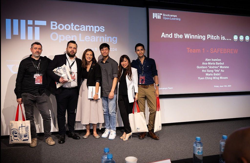 Group photo of 6 adults of different genders and races wearing business clothes posing in front of a screen with the logo for MIT Bootcamps at Open Learning and says, “And the winning pitch is…Team 1 SafeBrew.”