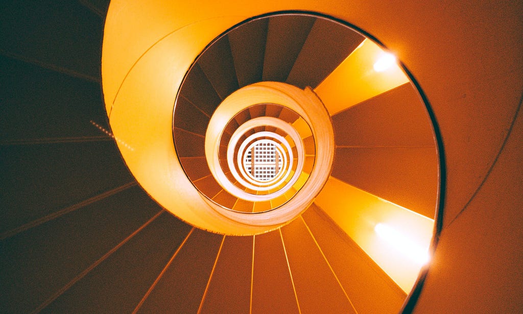 A spiral staircase viewed from below, along the middle of the spiral, with the underside of the steps resembling an upside-down staircase, warmly lit by pairs of lamps on the underside of steps on the left and right sides and leading up into a white grate.