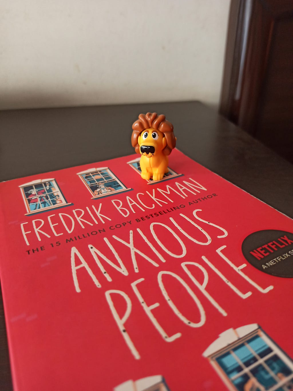 The book “Anxious People” by Fredrik Backman is placed in a table. The image shows a close up of the book. A small kinder egg lion toy is seated on top of the book.