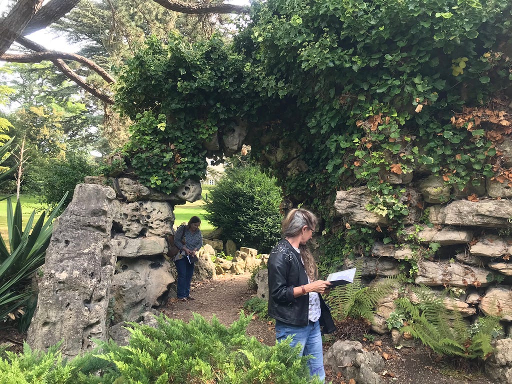 A woman is holding a notebook and she is looking sideways at stones — tufa stones which are grey, large, quite round and very holey. And the stones, they are balanced on each other to create an arch, and there are two low stone tufa walls either side of the woman who has a black jacket and fair hair and she is wearing jeans. And this is the Grotto from Sydney Gardens that was moved to the grounds of the Bath Spa Hotel.