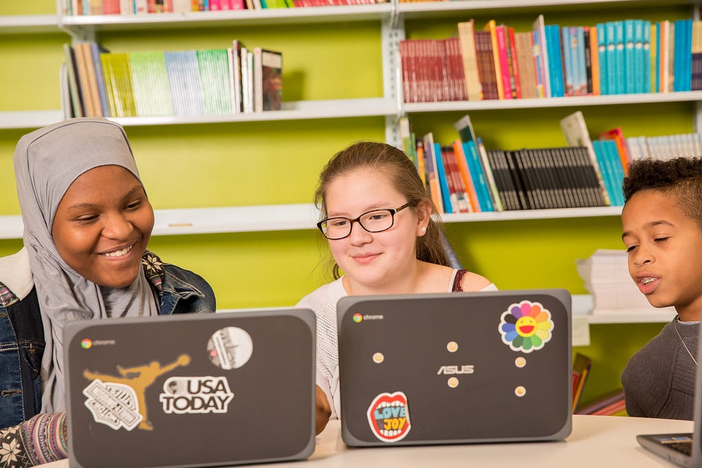 Three students looking at laptops and smiling in front of a bookcase.