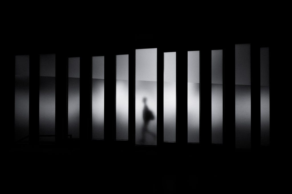 Silhouette of a blurred human carrying a bag framed in one pane while walking through a black and white field of jagged, slender, fogged, separate vertical panels which are surrounded in black