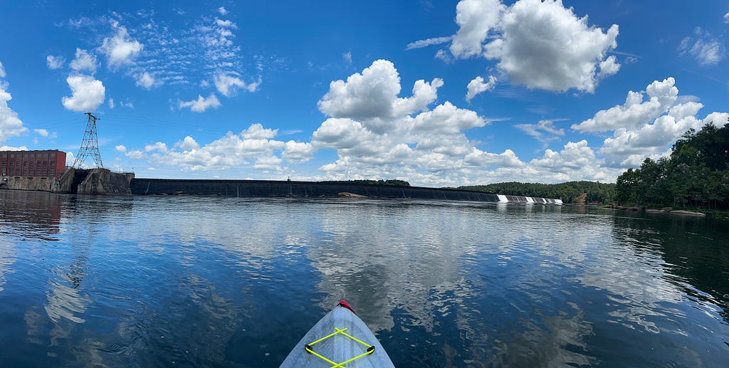 Panoramic view of entire dam — brick building on left with water flowing over the dam to the far right. Clear water all around.