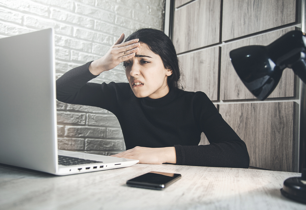 Woman looking frustrated with her right hand on her forehead. She is looking at a laptop. Wearing a black turtleneck. She has long black hair in a ponytail. There’s also a phone and a lamp next to her on a desk.