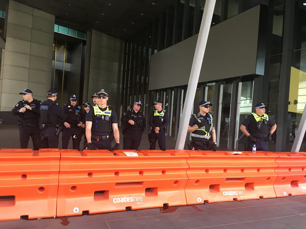 A row of bright orange barricades, some distance from the doors of a sleek structure, behind which cops wearing caps and vests stand clustered, many with sunglasses and thick gloves