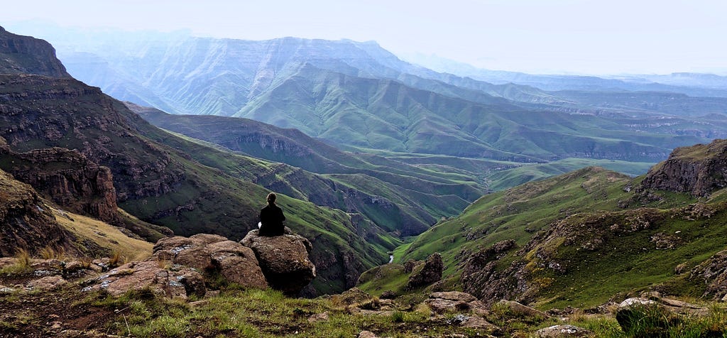 Women seen from behind sitting with crossed legs on rock while overlooking lush, green mountain valley